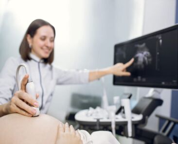 Cropped shot of a pregnant woman during ultrasound scanning at the fertility clinic. Female doctor pointing at the screen of ultrasound machine. Focus on ultrasound transducer on the pregnant belly.