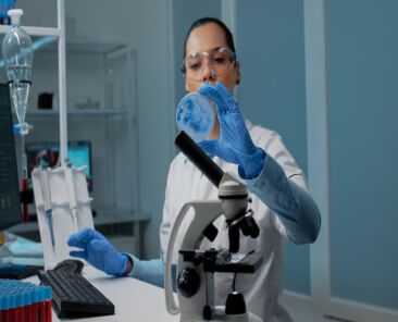 Chemistry scientist using research equipment in laboratory on desk with petri dish, micropipette, vacutainers and microscope. Scientific woman typing on computer for development